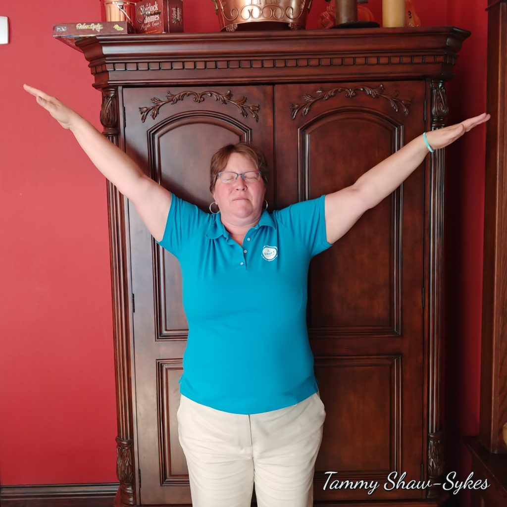 Tammy Shaw-Sykes standing in yoga pose with arms stretched over her head in front of wooden cabinet