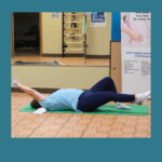 A person in gym attire is lying on an exercise mat on a wooden floor, performing a stretching exercise. They are holding a small weight in one hand, with their arm extended above their head, and their other arm and leg bent. A mirror and fitness equipment are in the background.